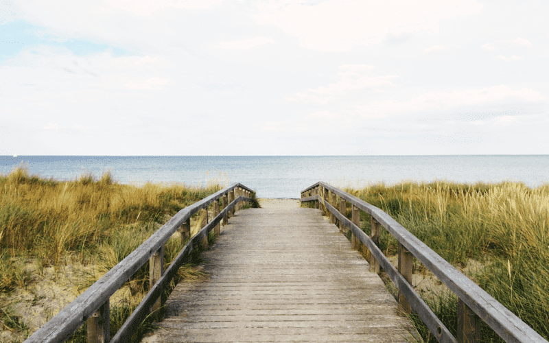 A wooden path leads through tall grass to a calm ocean under a cloudy sky.