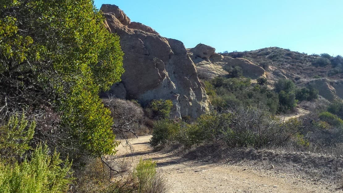 A winding dirt path surrounded by shrubs and rocky hills under a clear blue sky.