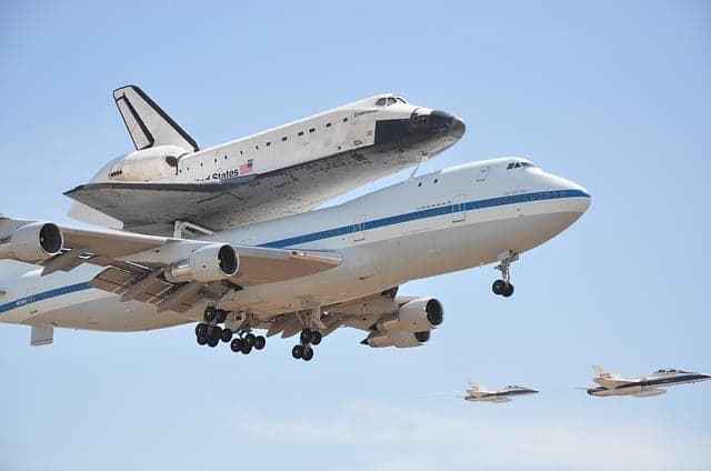 A space shuttle is mounted on top of a modified Boeing 747, with fighter jets flying in formation nearby.