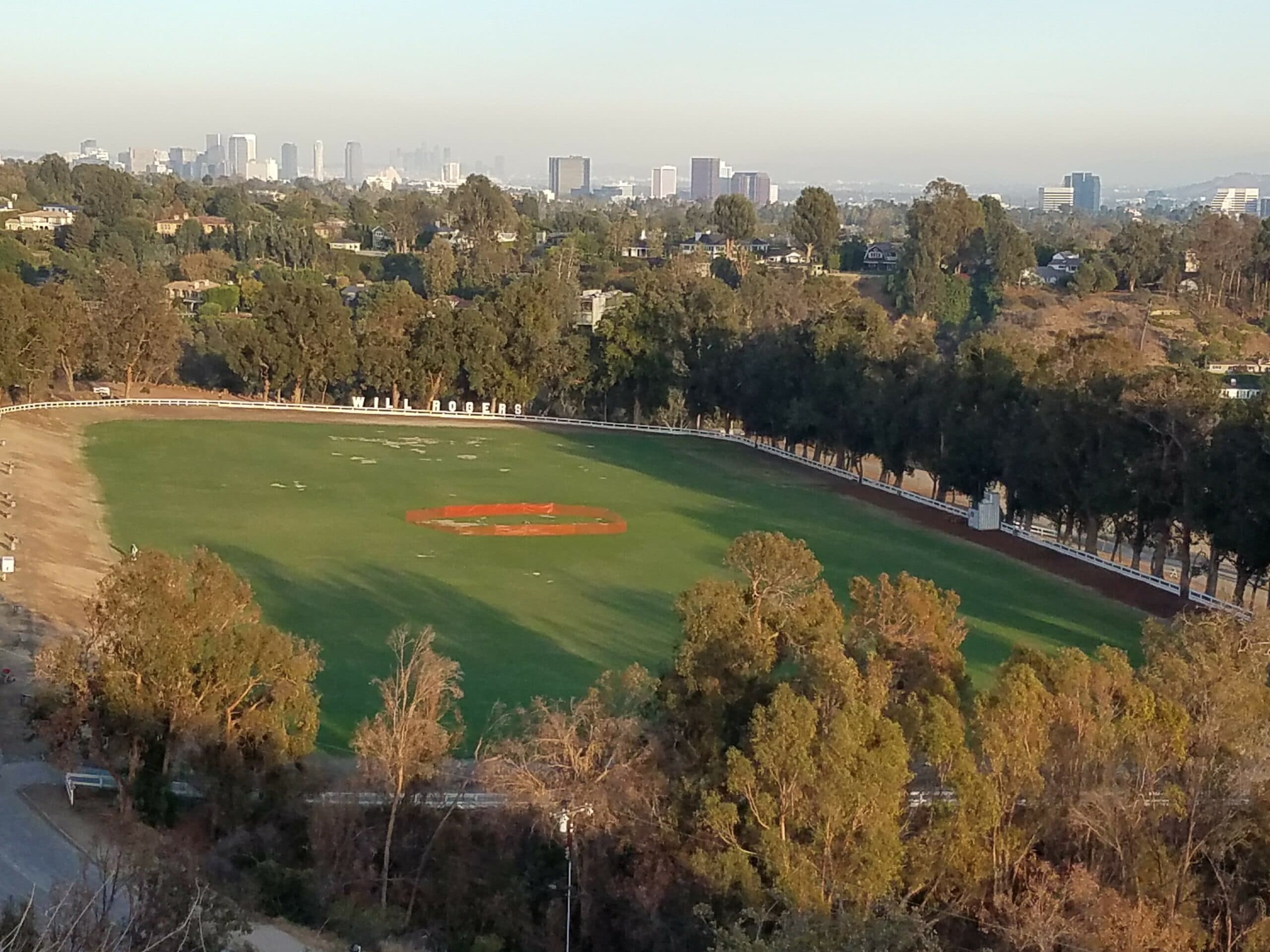 A panoramic view of a green sports field surrounded by trees, with a city skyline in the background.