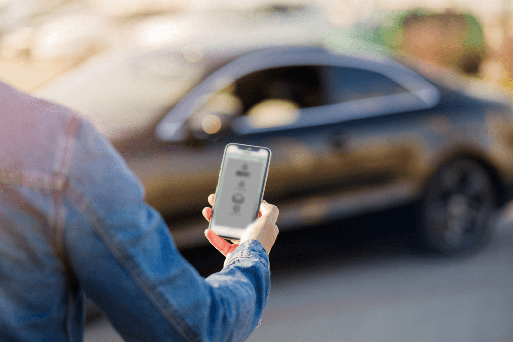 A person holds a smartphone while standing near a parked car.
