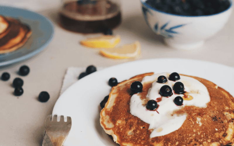 A plate of pancakes topped with yogurt and blueberries, accompanied by lemon slices and a bowl of blueberries in the background.