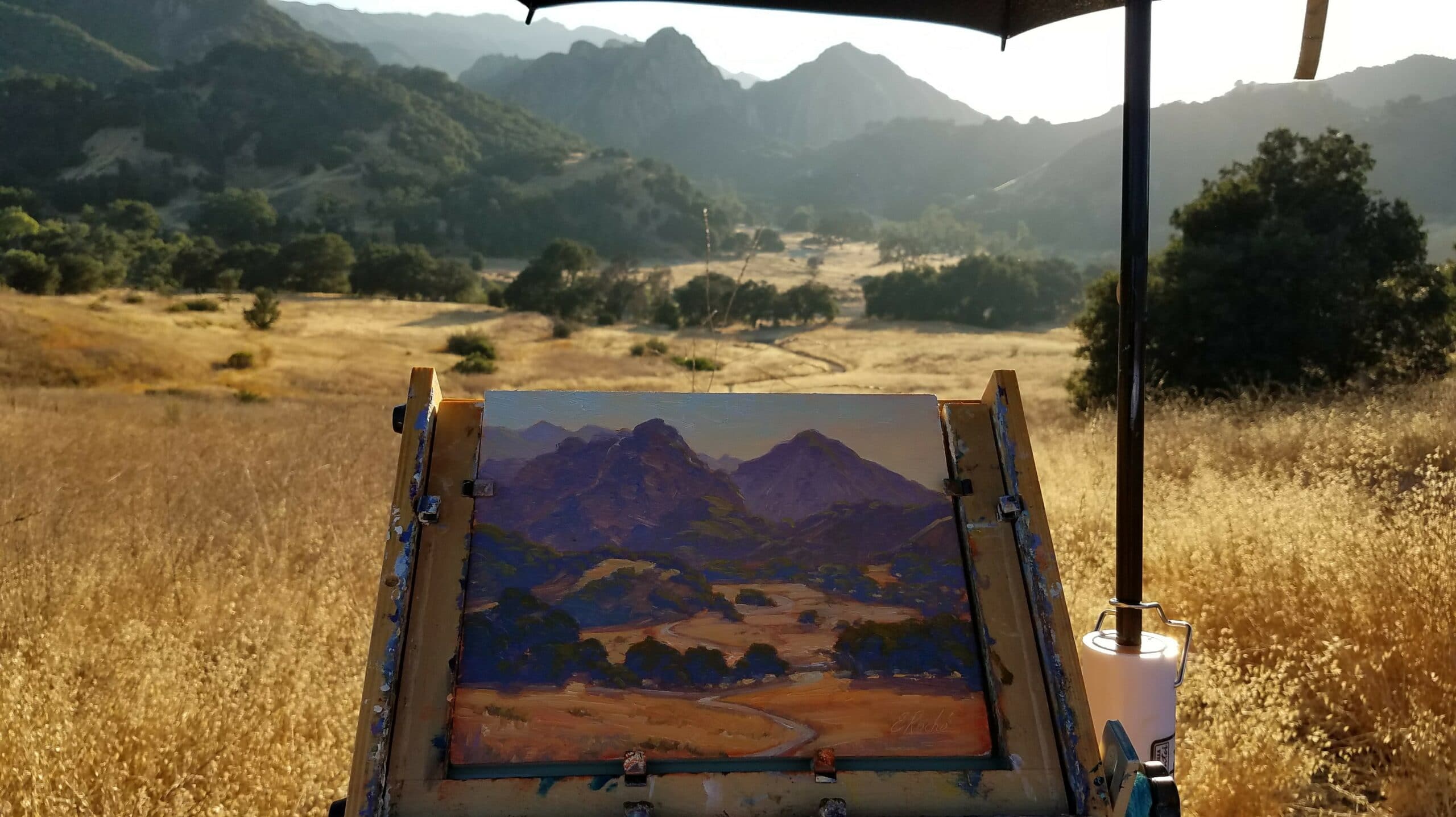 An easel displaying a painted landscape view of mountains and fields under a bright sky.