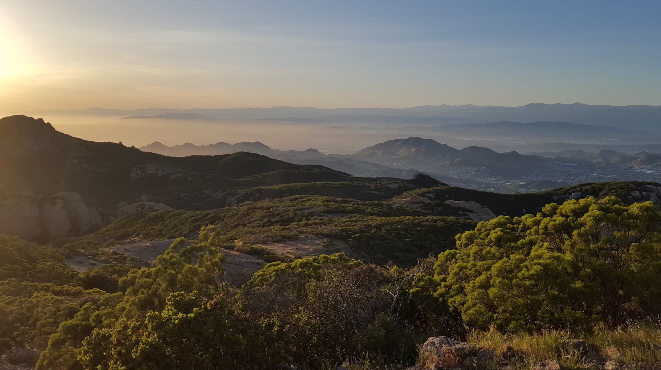 A panoramic view of misty mountains at sunset, framed by greenery in the foreground.