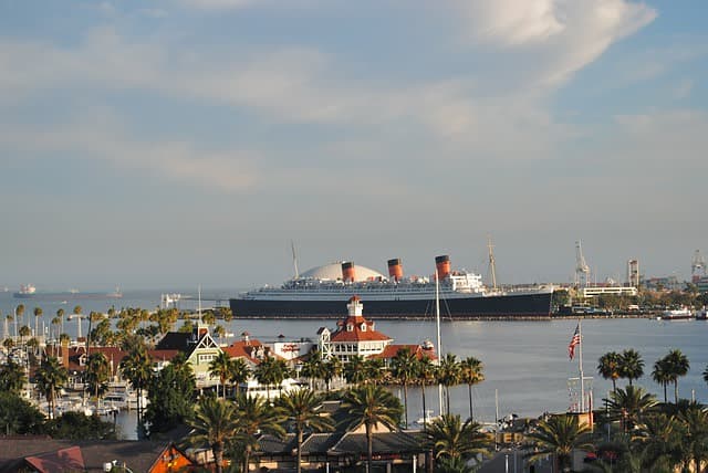 A historic ship is docked near palm-lined waterfront buildings under a cloudy sky.