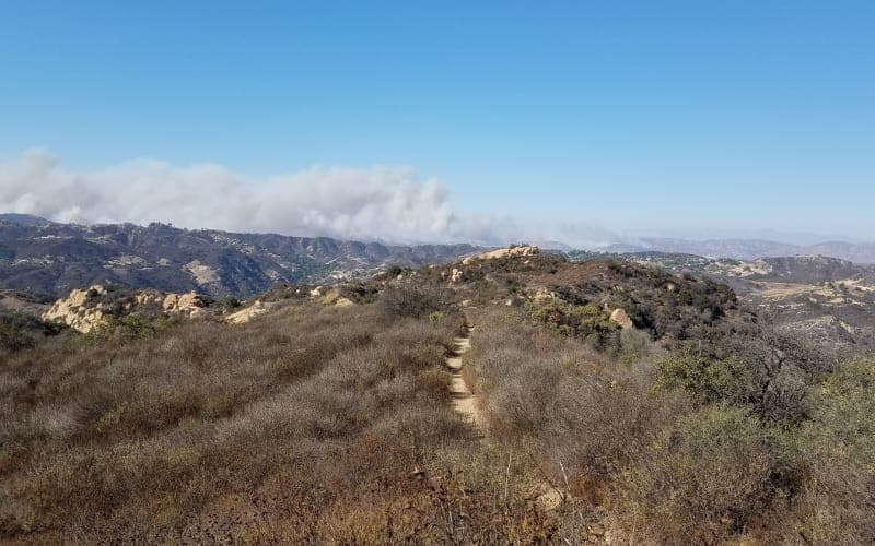 A dirt path winds through dry grasses and rocky terrain, with smoke billowing in the distance under a clear blue sky. A dirt path winds through dry grasses and rocky terrain, with smoke billowing in the distance under a clear blue sky.