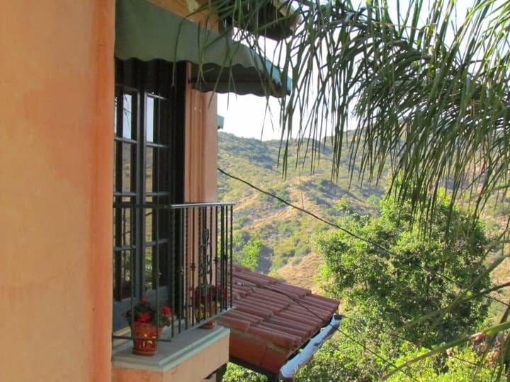 A balcony with potted flowers overlooks rolling green hills under a clear sky.