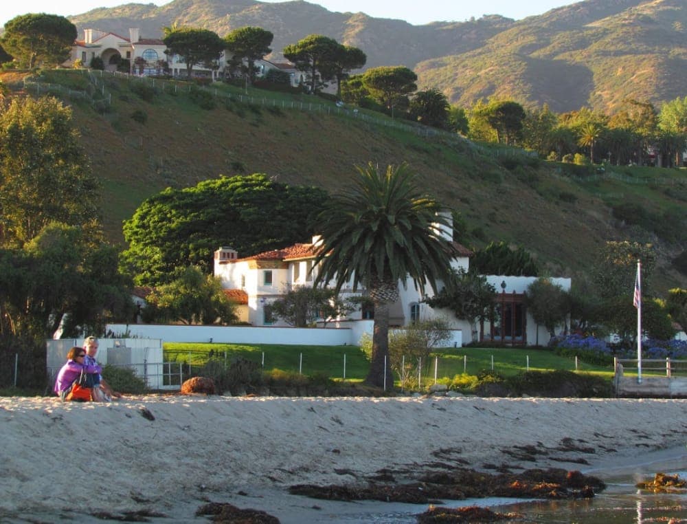 A sandy beach with two people sitting in front of a white house surrounded by lush greenery and hills. A sandy beach with two people sitting in front of a white house surrounded by lush greenery and hills.