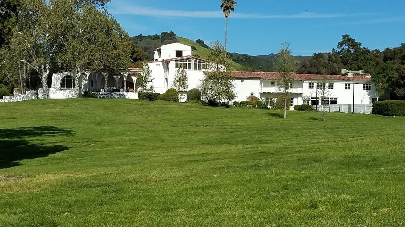 A white building with a red tile roof sits on a green grassy landscape, surrounded by trees and hills.