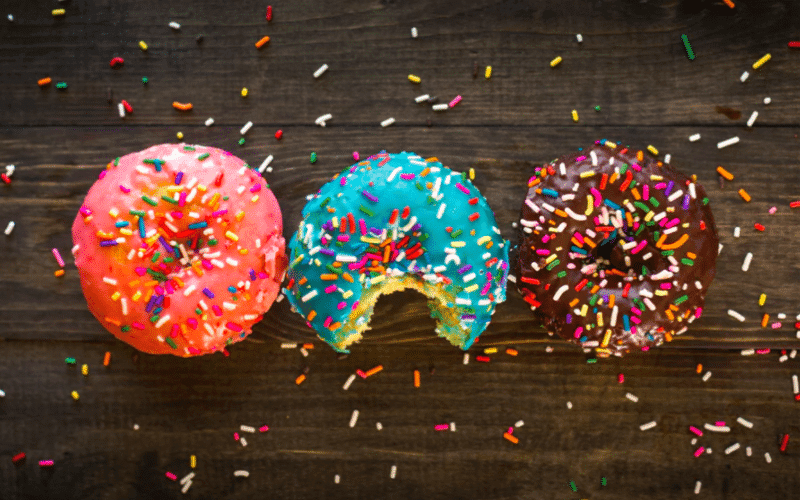 Three colorful donuts with sprinkles on a wooden surface, one partially eaten. Three colorful donuts with sprinkles on a wooden surface, one partially eaten.
