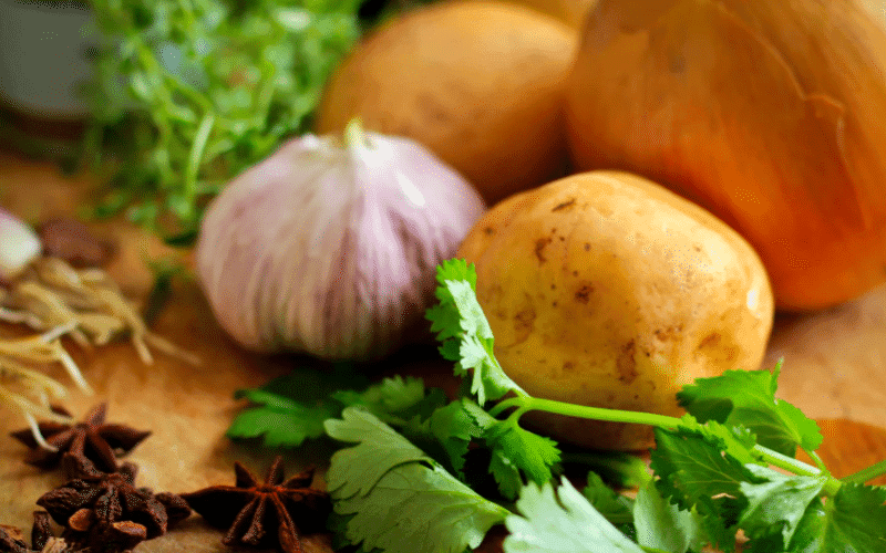 A collection of fresh vegetables, including garlic, potatoes, cilantro, and aromatic spices, arranged on a wooden surface. A collection of fresh vegetables, including garlic, potatoes, cilantro, and aromatic spices, arranged on a wooden surface.