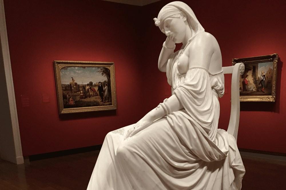 A white marble statue of a seated woman in a contemplative pose, with paintings on the wall behind her.