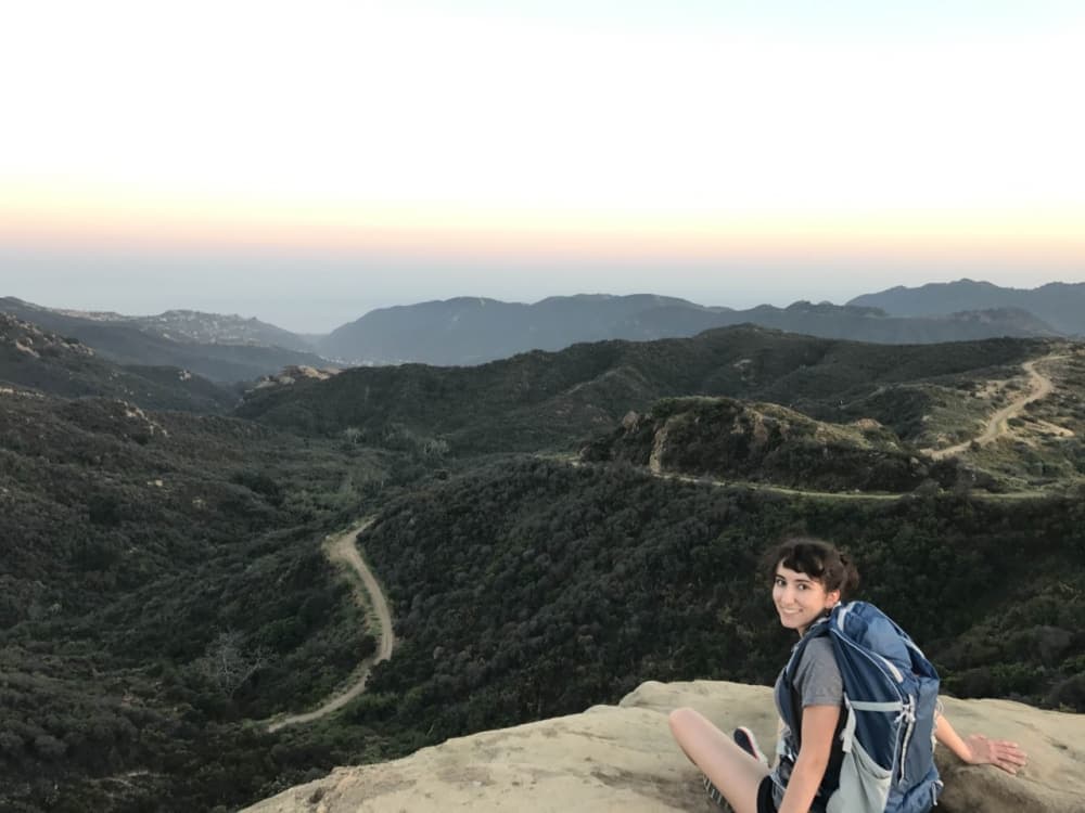 A person sitting on a rock ledge overlooking a scenic mountainous landscape at sunset.