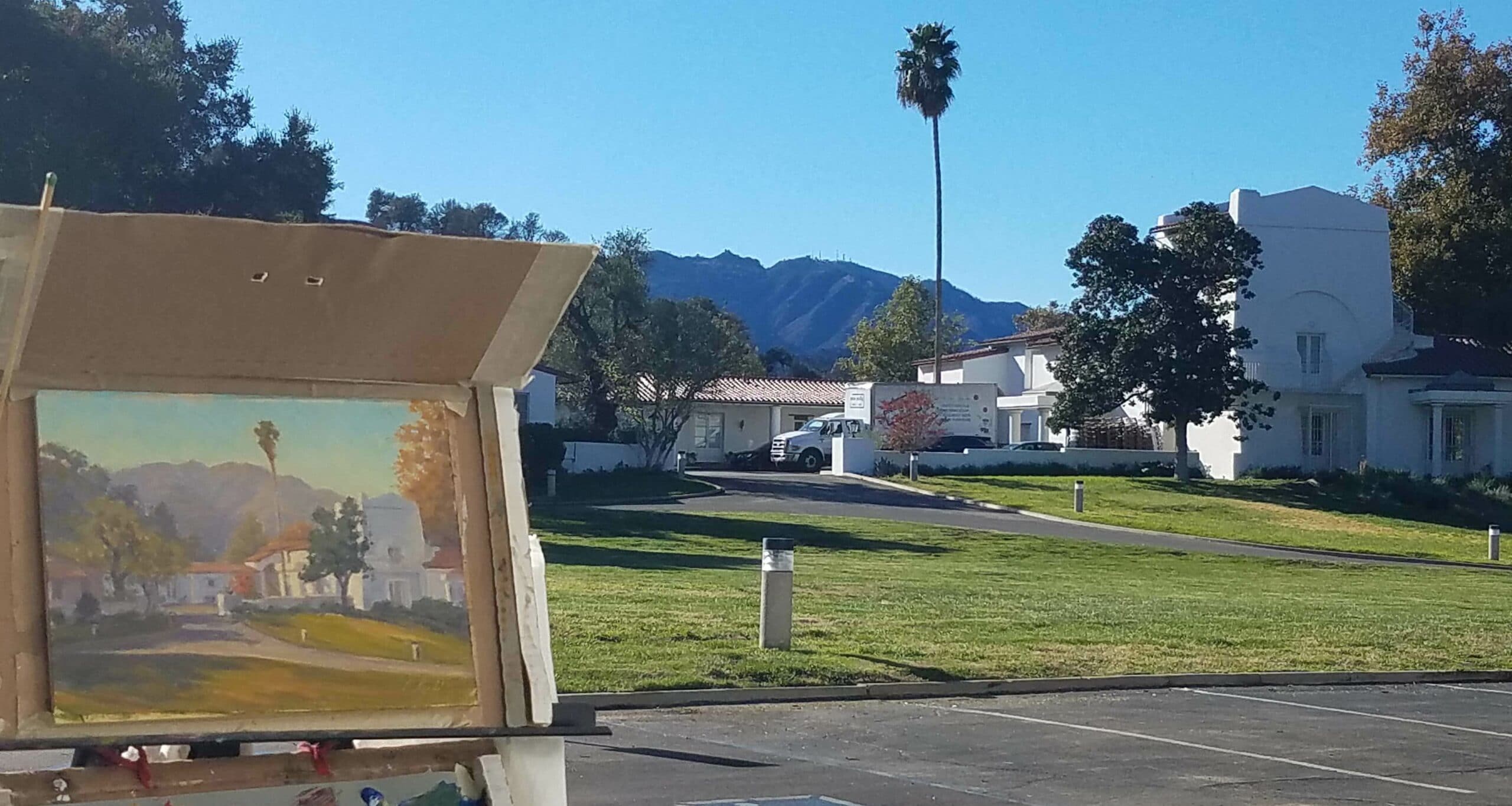 An easel with a landscape painting in the foreground, showcasing a sunny view of houses and mountains in the background.