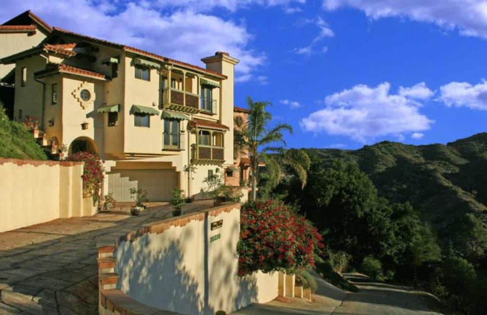 A picturesque hillside house with a curved driveway, surrounded by greenery and under a blue sky.