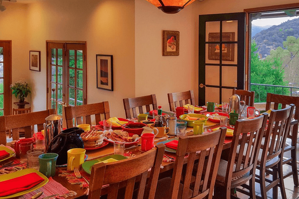 A dining table set with colorful dishes and drinks, surrounded by chairs and open doors leading to a scenic view.
