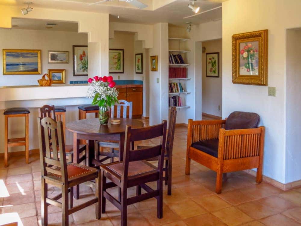 A bright dining area featuring a wooden table, chairs, and floral arrangements, with shelves of books and artwork on the walls.